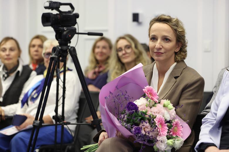 Kobieta siedzi na krześle na sali konferencyjnej trzymając bukiet kwiatów