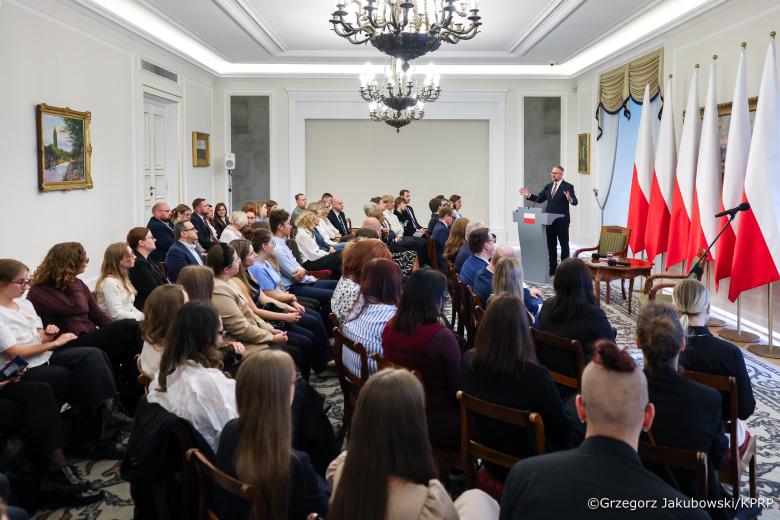 Kilkadziesiąt osób siedzi na sali i słucha wystąpienia osoby mówiącej za mównicą