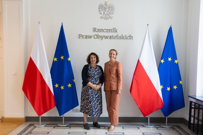 Dwie osoby pozują na stojąco do wspólnego zdjęcia na tle ściany z napisem "Rzecznik Praw Obywatelskich" i flag Polski i UE