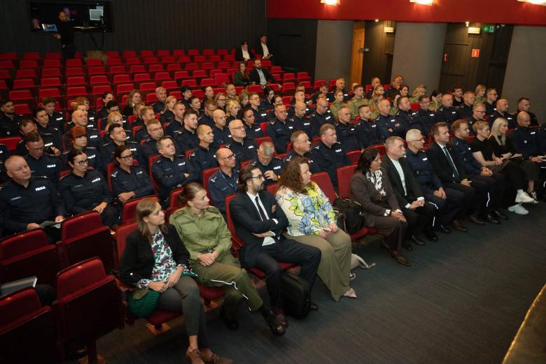 Kilkadziesiąt osób siedzi w fotelach na widowni w teatrze i słucha wystąpienia