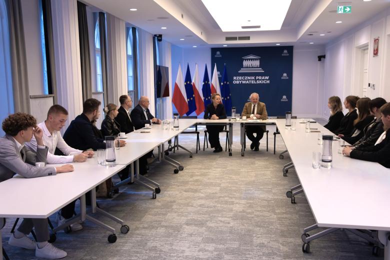 Kilkanaście osób siedzi przy stołach ustawionych w podkowę na sali konferencyjnej