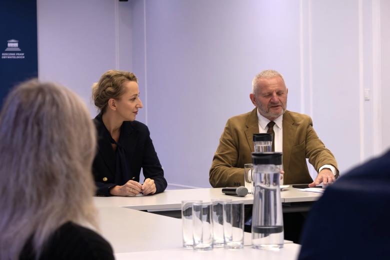 Dwie osoby siedzą za stołem na sali konferencyjnej