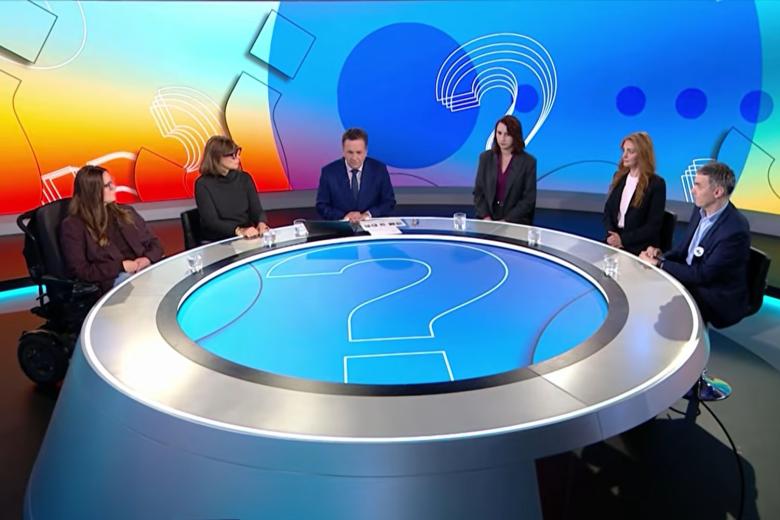 Zrzut ekranu z transmisją programu telewizyjnego. Sześć osób siedzi przy stole w studio telewizyjnym i rozmawia