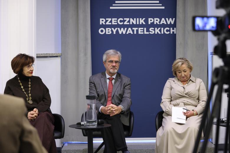 Trzy osoby siedzą na krzesłach w szeregu w sali konferencyjnej