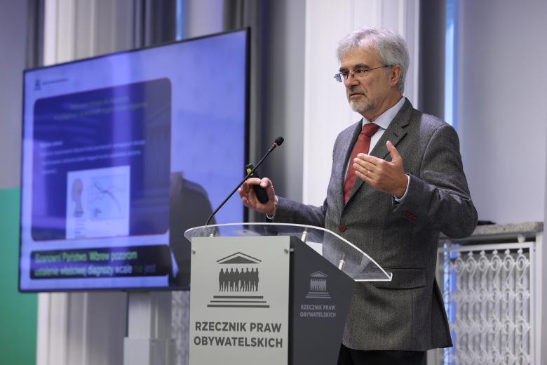 Mężczyzna stoi przy mównicy i wypowiada się, gestykulując. Za nim widać ekran wyświetlający prezentację