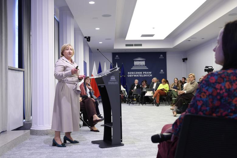Kobieta wypowiada się, stojąc przy mównicy w sali konferencyjnej. Naprzeciwko niej na widowni siedzi kilkadziesiąt osób