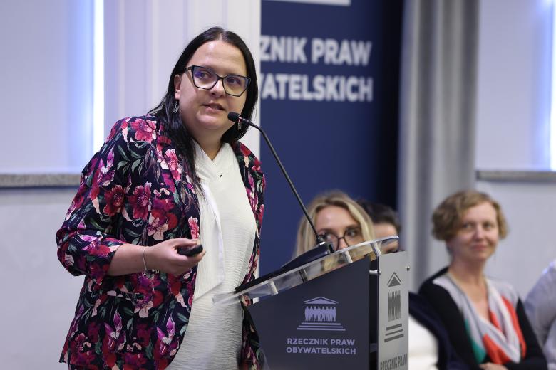 Kobieta stoi przy mównicy i wypowiada się, patrząc na obiekt poza kadrem. W tle za nią widać trzy osoby siedzące w panelu dyskusyjnym