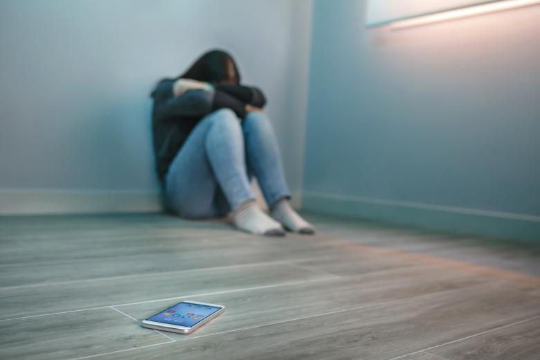 Skulona kobieta z głową schowaną w ramionach siedzi pod ścianą na podłodze pustego pokoju, przed nią na podłodze leży włączony telefon