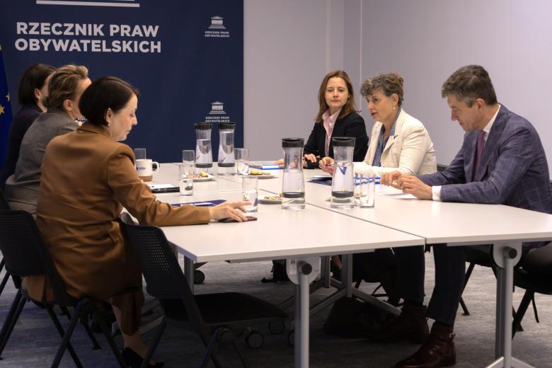 Sześć osób siedzi po dwóch stronach dużego stołu w sali konferencyjnej i prowadzi rozmowę