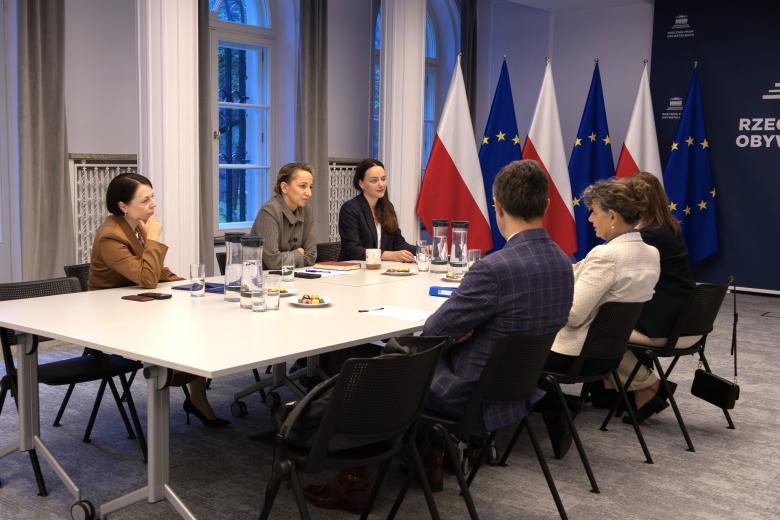 Sześć osób siedzi po dwóch stronach dużego stołu w sali konferencyjnej i prowadzi rozmowę