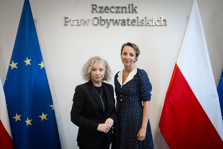 Sylwia Gregorczyk-Abram i Katarzyna Kotula uśmiechają się pozując do zdjęcia na tle flag Polski i Unii Europejskiej oraz napisu "Rzecznik Praw Obywatelskich"