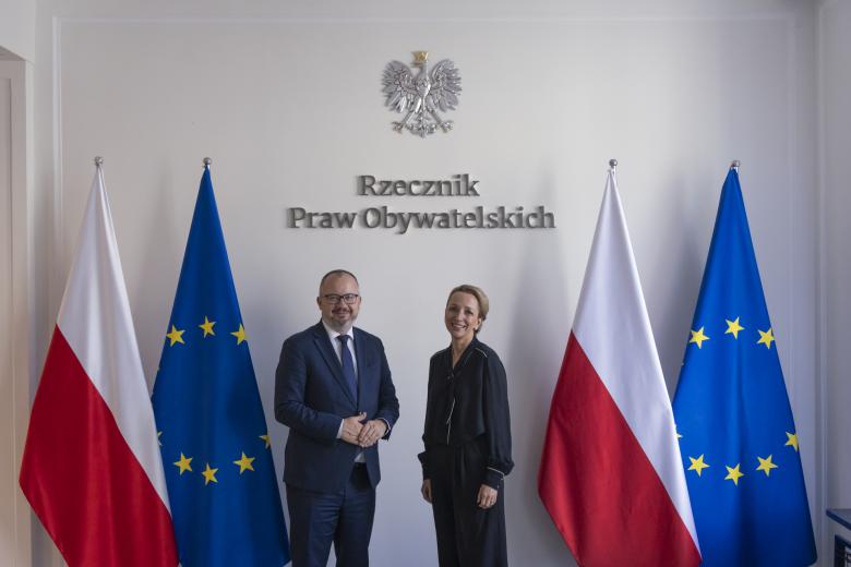 Sylwia Gregorczyk-Abram i Adam Bodnar pozują wspólnie do zdjęcia, uśmiechając się na tle flag Polski i Unii Europejskiej, godła Polski i napisu "Rzecznik Praw Obywatelskich"