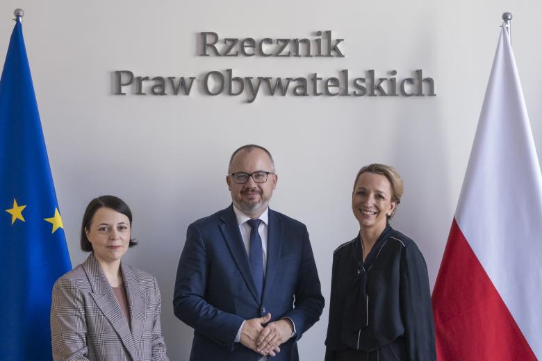 Sylwia Gregorczyk Abram, Anna Błaszczak-Banasiak i Adam Bodnar pozują do zdjęcia uśmiechając się na tle flag Polski i Unii Europejskiej i napisu "Rzecznik Praw Obywatelskich"