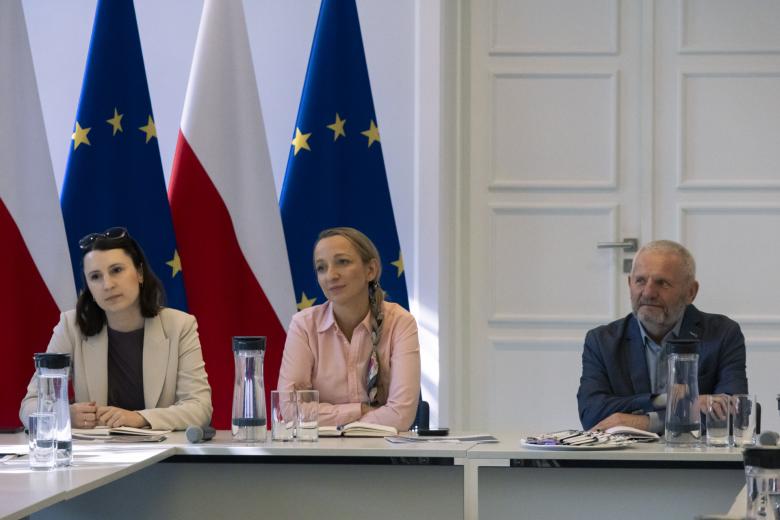 Trzy osoby siedzą przy stole konferencyjnym. W tle flagi Polski i Unii Europejskiej