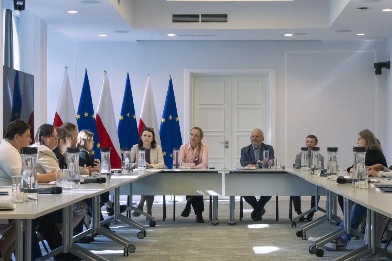Kilkanaście osób rozmawia siedząc przy stole konferencyjnym w kształcie podkowy. W tle flagi Polski i Unii Europejskiej