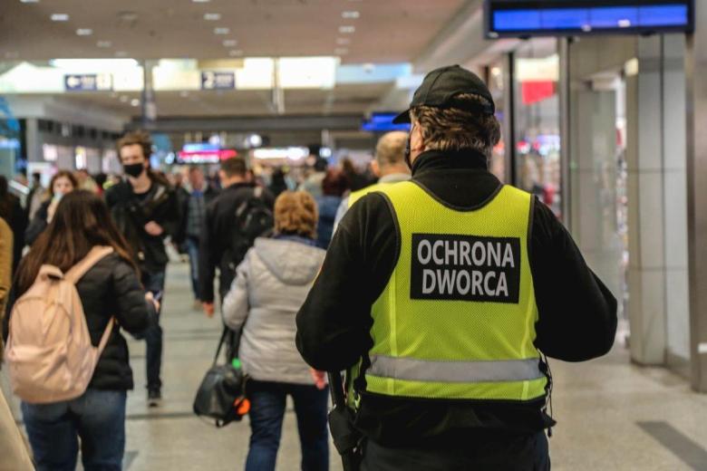 Osoba w mundurze i zielonej kamizelce z napisem "Ochrona dworca" idzie przez zatłoczony korytarz podziemny