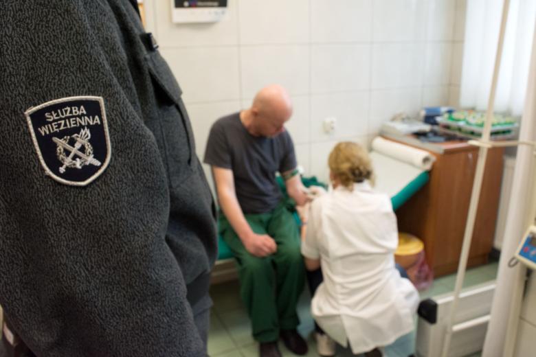 Pielęgniarka rozmawia z osobą siedząca na leżance w ambulatorium, na pierwszym planie ramie osoby w mundurze z naszywką z napisem "Służba Więzienna"
