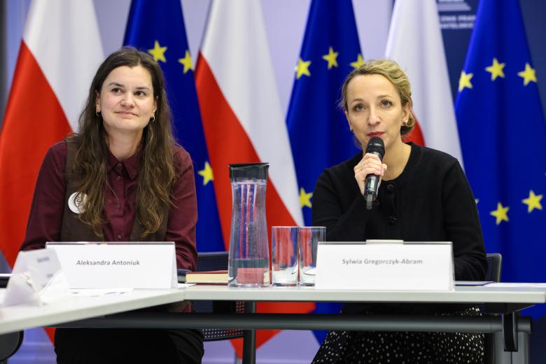 Dwie osoby, od prawej przez mikrofon mówi RPO Sylwia Gregorczyk-Abram, w tle flagi Polski i UE