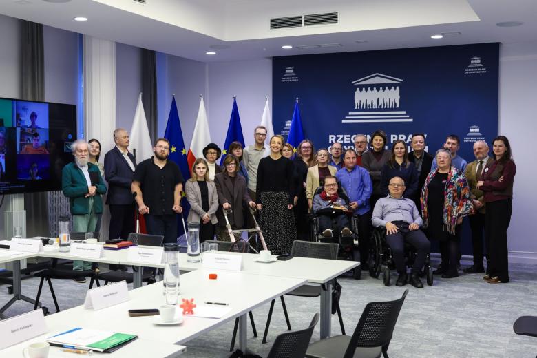 Kilkanaście osób pozuje na stojąco do zdjęcia na tle flag Polski i UE