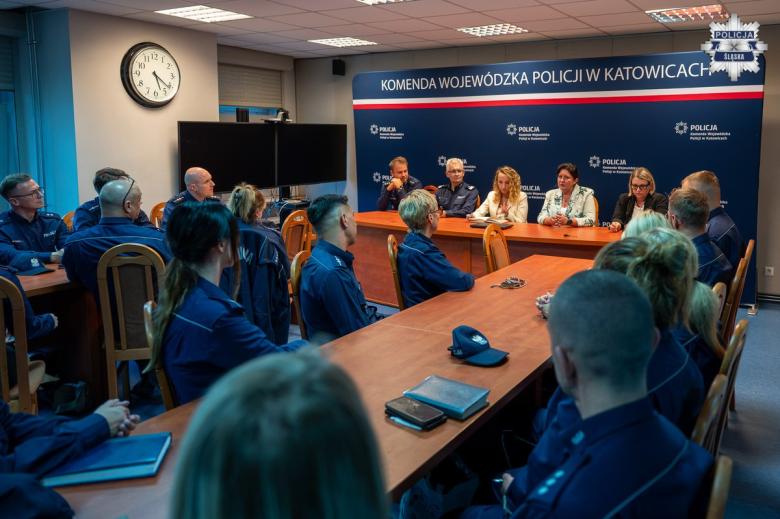 Kilkanaście osób w mundurach policyjnych siedzi na salce konferencyjnej, przed nimi pięć osób siedzi za stołem na tle dużej planszy z napisem "Komenda Wojewódzka Policji w Katowicach"
