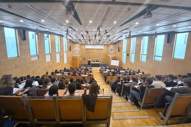 Kilkadziesiąt osób siedzi na dużej, uniwersyteckiej auli wykładowej i słucha wystąpienia osoby mówiącej na scenie za mównicą
