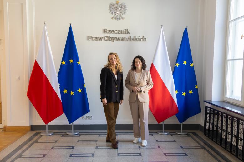 Dwie osoby pozują na stojąco do wspólnego zdjęcia na tle ściany z napisem "Rzecznik Praw Obywatelskich" i flag Polski i UE