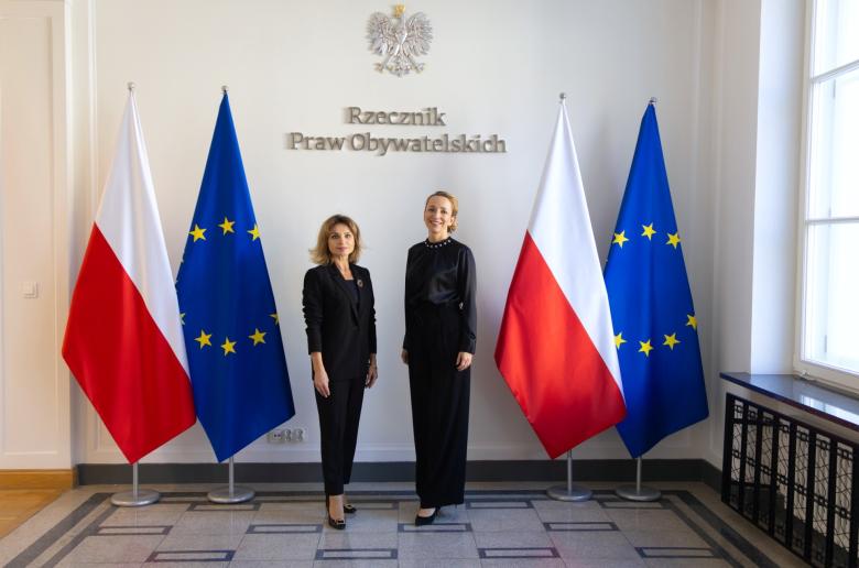 Dwie osoby pozują na stojąco do wspólnego zdjęcia na tle ściany z napisem "Rzecznik Praw Obywatelskich" i flag Polski i UE