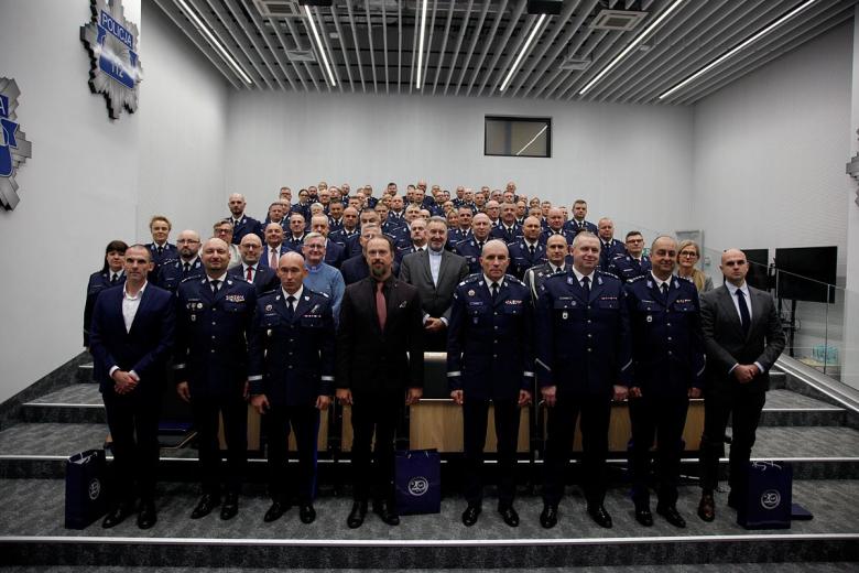 Kilkadziesiąt osób pozuje na stojąco do wspólnego zdjęcia na sali wykładowej