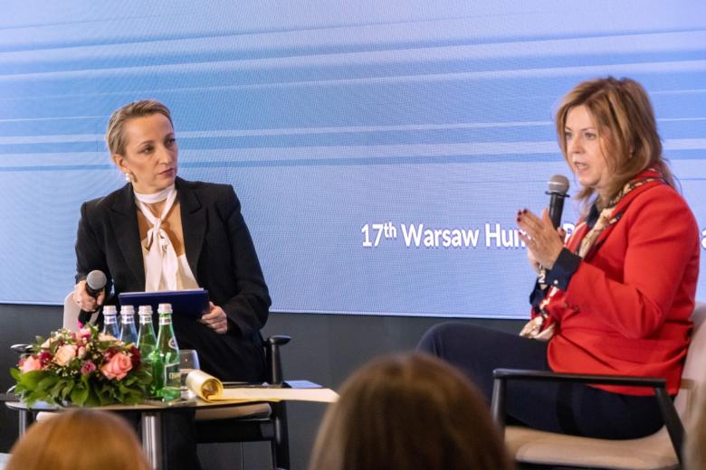 Dwie osoby siedzą na krzesłach na scenie i rozmawiają trzymając mikrofony