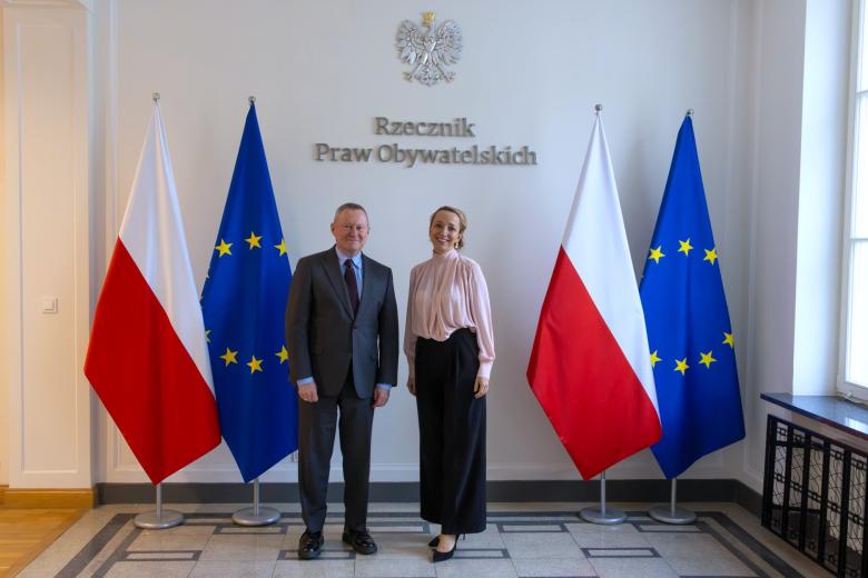 Dwie osoby pozują na stojąco do wspólnego zdjęcia na tle ściany z napisem "Rzecznik Praw Obywatelskich" i flag Polski i UE