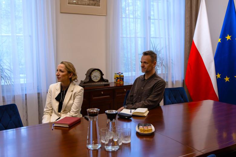 Dwie osoby siedzą przy stole i słuchają wypowiedzi osoby połączonej zdalnie na spotkanie