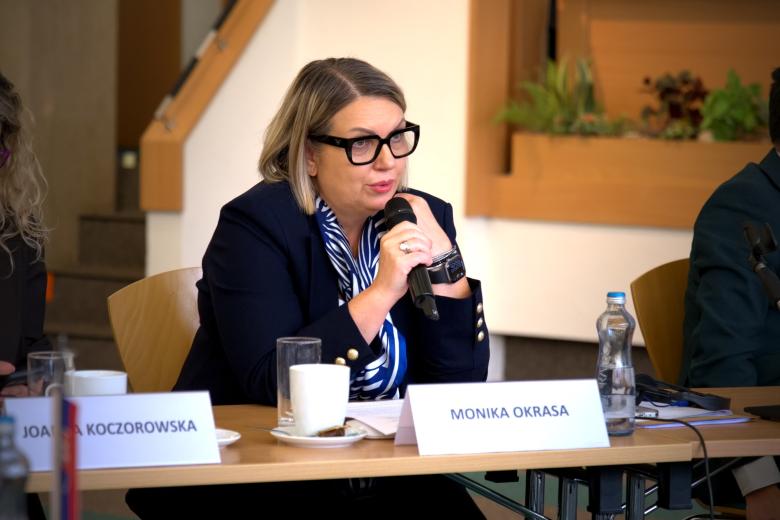 Osoba siedzi za stołem i mówi do trzymanego w dłoni mikrofonu