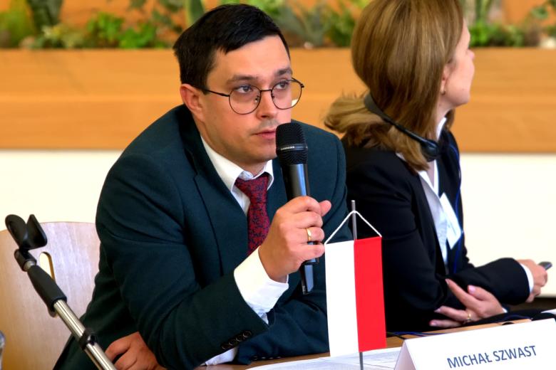 Osoba siedzi za stołem i mówi do trzymanego w dłoni mikrofonu