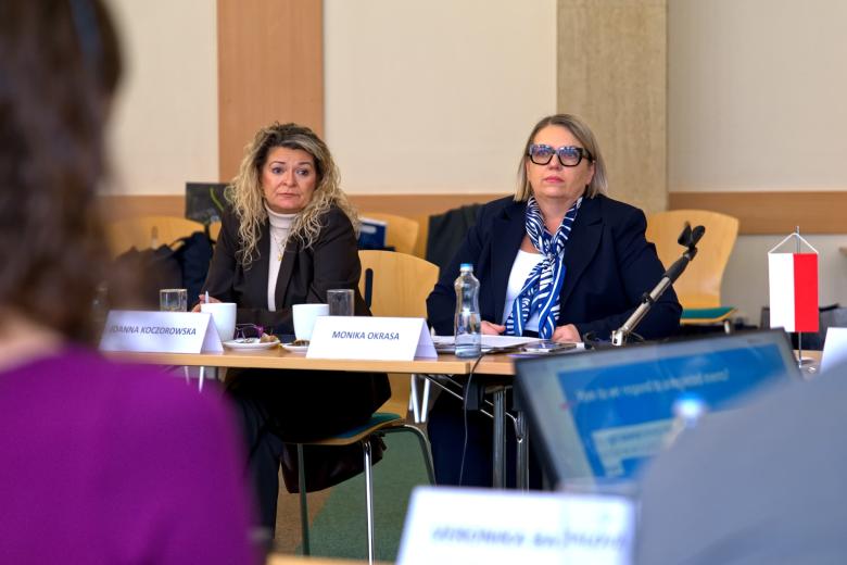 Dwie osoby siedzą za stołem na sali konferencyjnej, na stole mała, biało-czerwona flaga Polski