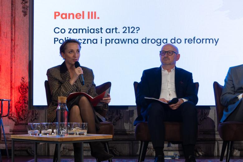 Dwie osoby siedzą na fotelach na scenie, jedna z nich mówi do trzymanego w dłoni mikrofonu