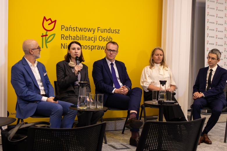 Pięć osób siedzi na krzesłach na sali konferencyjnej, jedna z nich mówi do trzymanego w dłoni mikrofonu