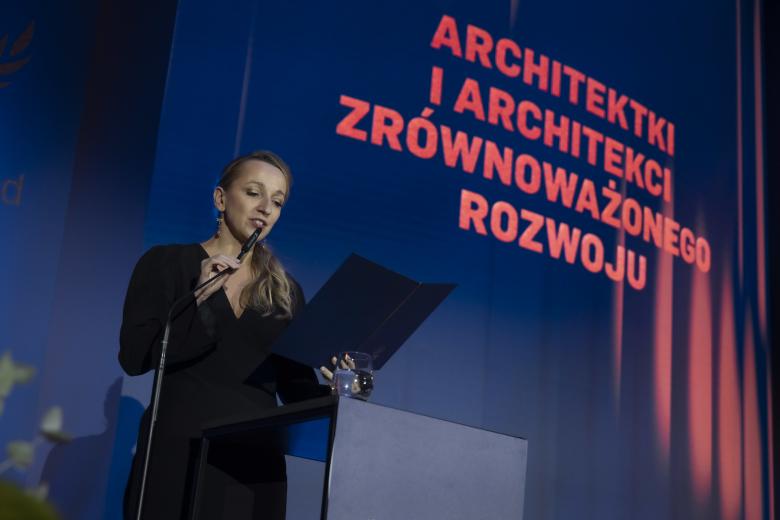 Osoba przemawia stojąc za mównicą na tle dużej planszy z napisem "Architektki i Architekci Zrównoważonego Rozwoju"