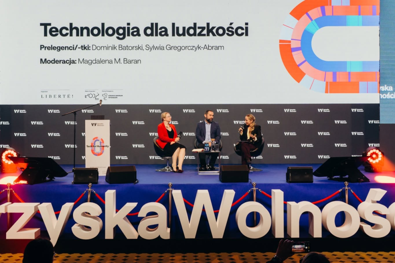 Zbliżenie na trzy osoby, które siedzą na fotelach na dużej scenie przed którą widoczny jest duży napis "Igrzyska Wolności", w tle na dużym wyświetlaczu zbliżenie na jedną z osób i grafika z napisem "Technologia dla ludzkości"