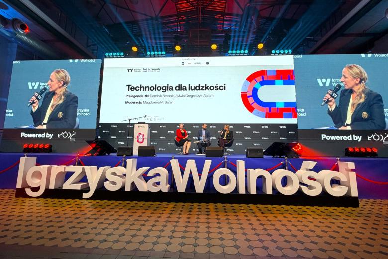 Trzy osoby siedzą na fotelach na dużej scenie przed którą widoczny jest duży napis "Igrzyska Wolności", w tle na dużym wyświetlaczu zbliżenie na jedną z osób i grafika z napisem "Technologia dla ludzkości"