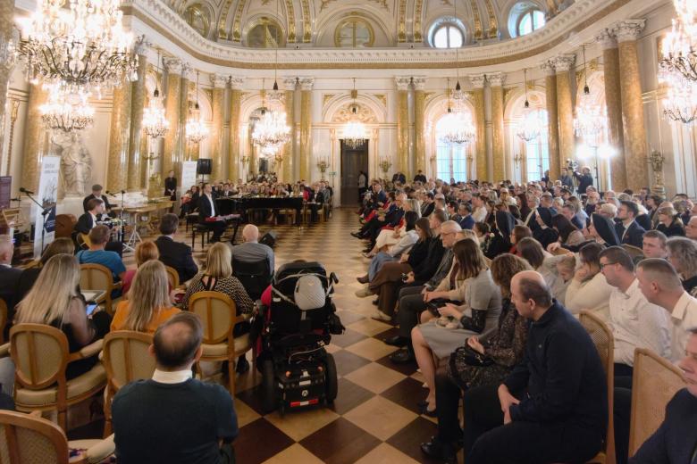 Bogato zdobiona, duża zabytkowa sala wypełniona ludźmi siedzącymi na krzesłach i słuchającymi występu muzycznego