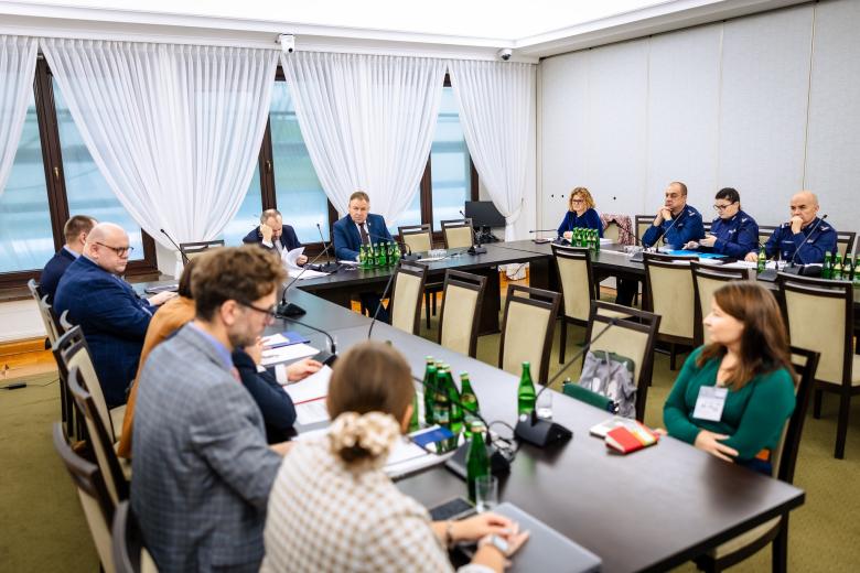 Kilkanaście osób siedzi przy stołach ustawionych w podkowę na sali konferencyjnej