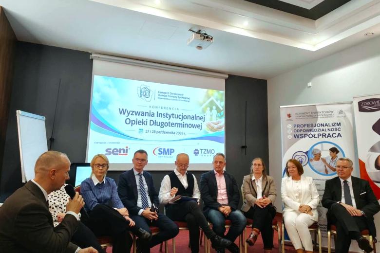 Dziewięć osób siedzi na krzesłach w sali konferencyjnej i słucha wypowiedzi jednej z nich mówiącej do mikrofonu