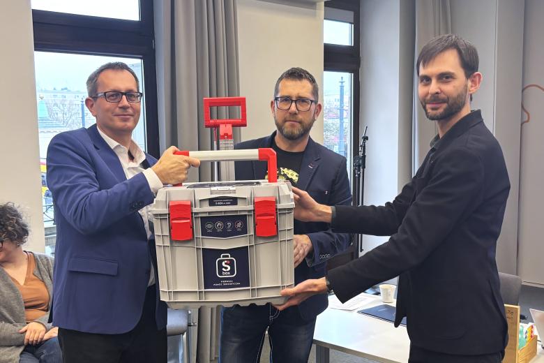 Trzy osoby pozują do wspólnego zdjęcia trzymając dużą skrzynkę z widocznym napisem "SBOX"