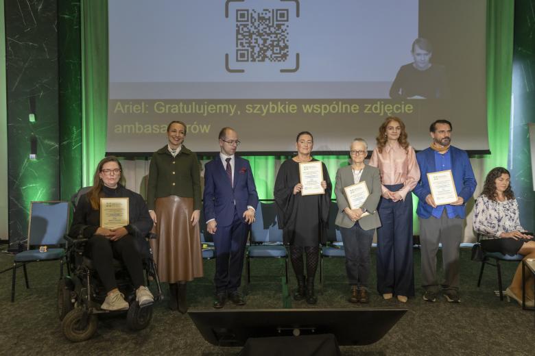 Osiem osób pozuje do wspólnego zdjęcia, niektóre z nich trzymają w rękach oprawione dyplomy, w tym jedna siedząca na wózku