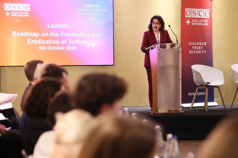 Osoba przemawia za mównicą na sali konferencyjnej, publiczność słucha wystąpienia