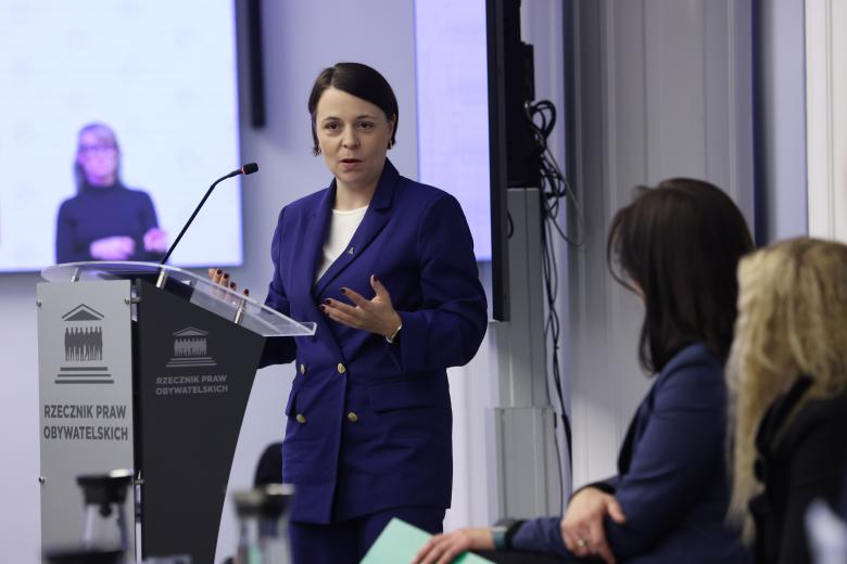 Kobieta w granatowej, dwurzędowej marynarce wypowiada się stojąc przy mównicy z mikrofonem i gestykuluje. Na pierwszym planie są sylwetki siedzących osób