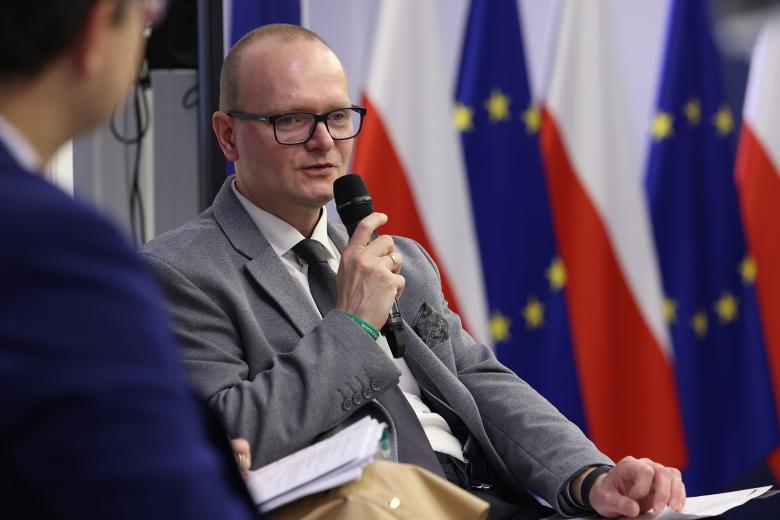 Mężczyzna w garniturze siedzi i wypowiada się do trzymanego w dłoni mikrofonu.