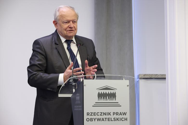Mężczyzna stoi przy mównicy i wypowiada się do mikrofonu, gestykulując dłońmi