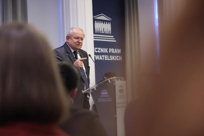Mężczyzna stoi przy mównicy i wypowiada się, gestykulując. Na pierwszym planie są rozmyte sylwetki osób z widowni