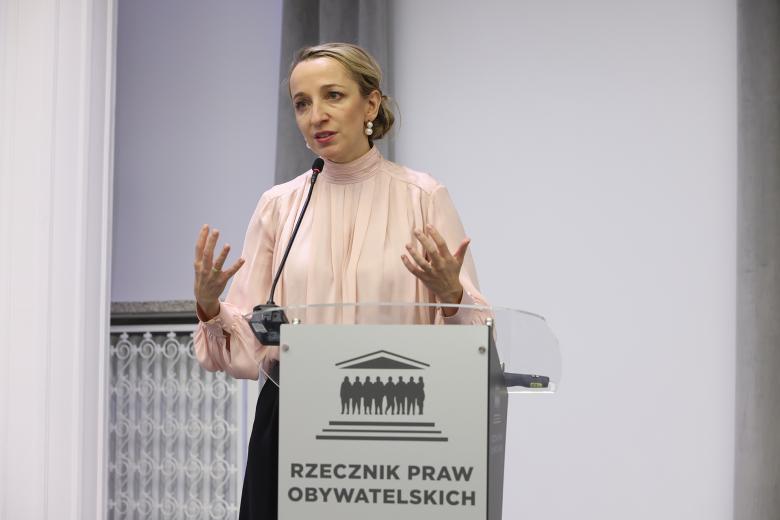 Kobieta w beżowej bluzce stoi przy mównicy i wypowiada się, gestykulując dłońmi